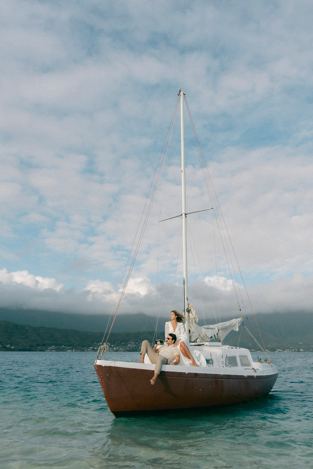  A couple lounging on a boat in the water, with the woman sitting at the mast and the man reclining at the front, enjoying the scenic view of the mountains and the sky.