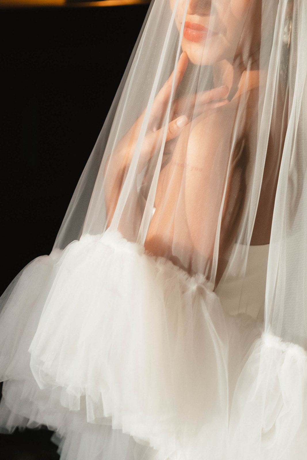 A dreamy close-up of a bride’s hands delicately touching her veil, with soft ruffled sleeves peeking through. The image captures a romantic and ethereal bridal moment.