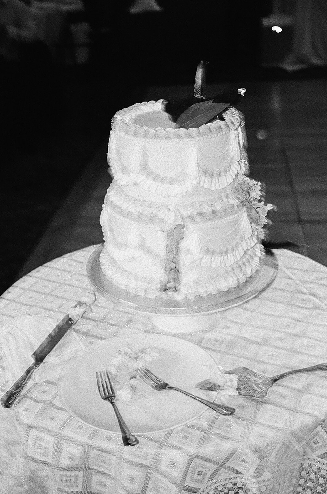 A vintage black and white photo of a wedding cake with intricate lace-like frosting details, slightly tilted on a table.