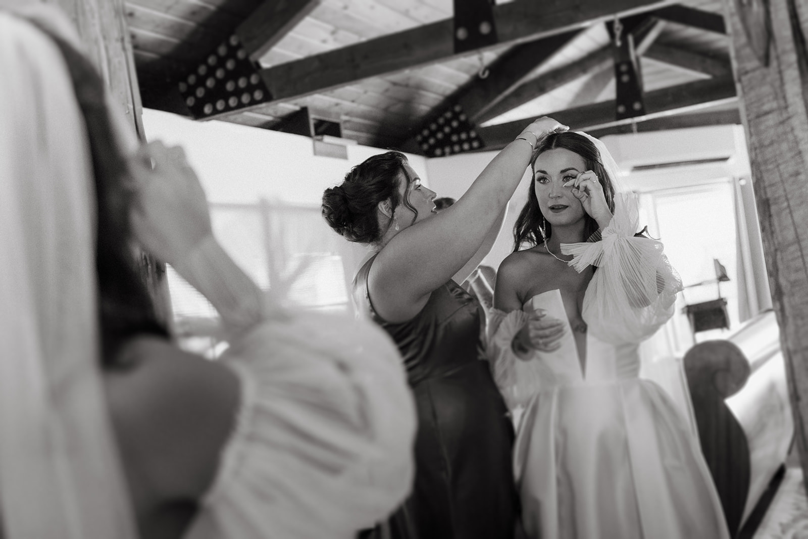 A black-and-white photo of a bride being helped by her bridesmaid as she adjusts her veil. The bride is in a white dress with puffed sleeves, while the bridesmaid, wearing a satin dress, carefully adjusts the bride's hair.