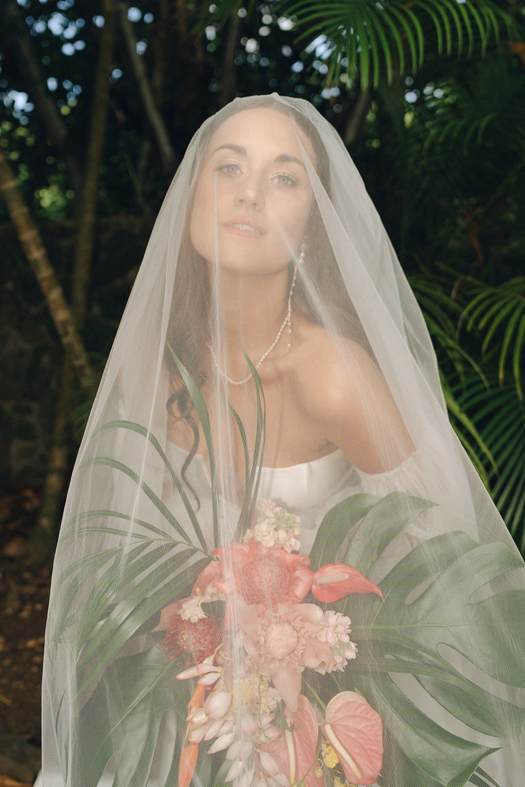 A bride stands under a delicate veil, holding a vibrant tropical bouquet with large palm leaves and pink anthuriums. The soft, diffused light highlights her serene expression against a lush green backdrop.