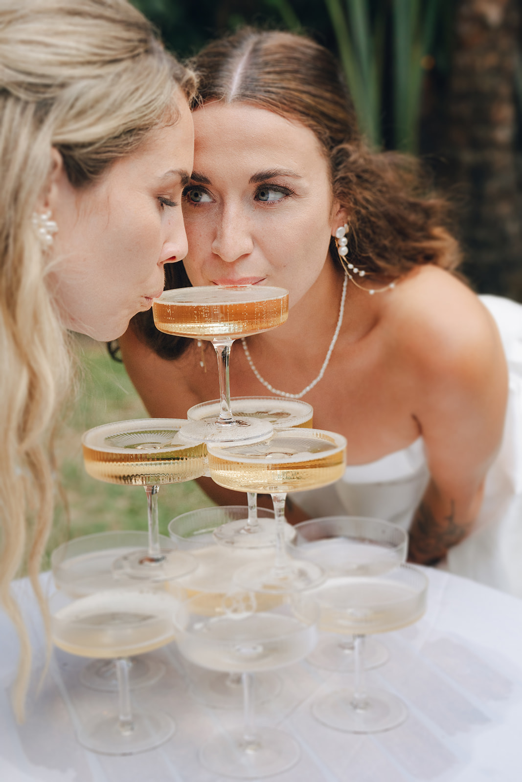 A bride and her bridesmaid lean in to sip from a champagne tower, perfectly capturing the celebratory mood of the wedding day. Golden bubbles and elegant coupe glasses add a luxe touch.