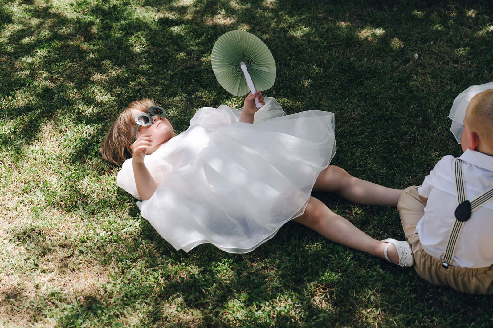 A flower girl in a white tulle dress lounges on the grass, sunglasses on and paper fan in hand. The ultimate wedding day mood: effortless and unbothered.