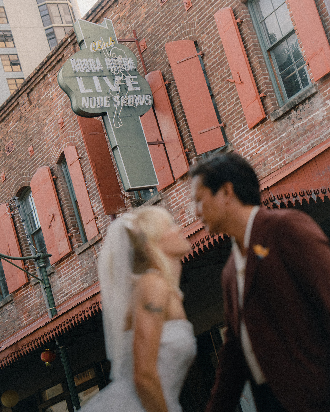 A blurred photo of a bride and groom standing in front of a brick building with a neon sign that reads "Live Nude Shows" above them. The bride is in a white wedding dress with a veil, while the groom is wearing a burgundy suit.