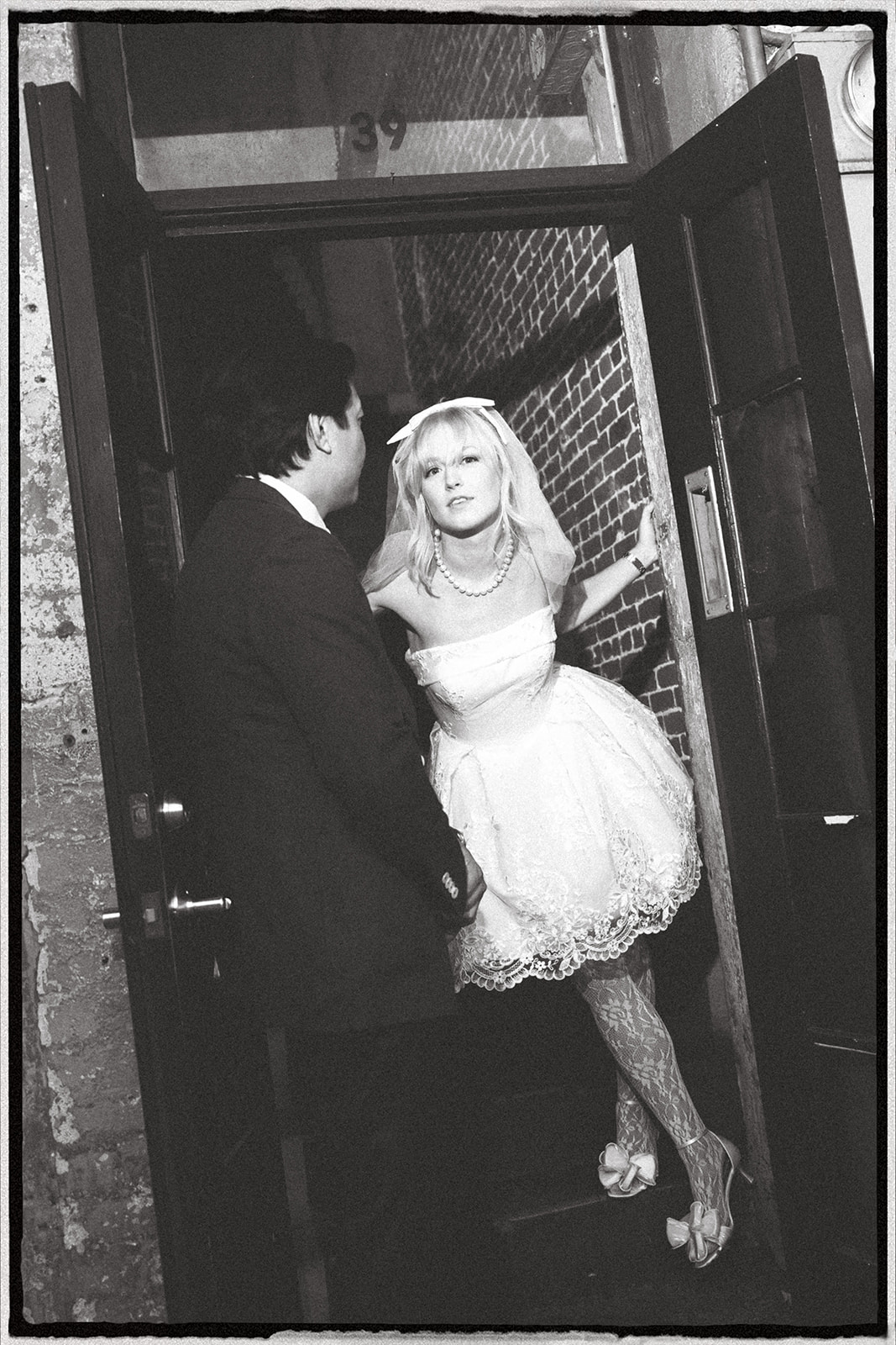A bride in a short white dress with a veil and pearl necklace stands at a door, looking at her partner, while he stands by the doorframe.