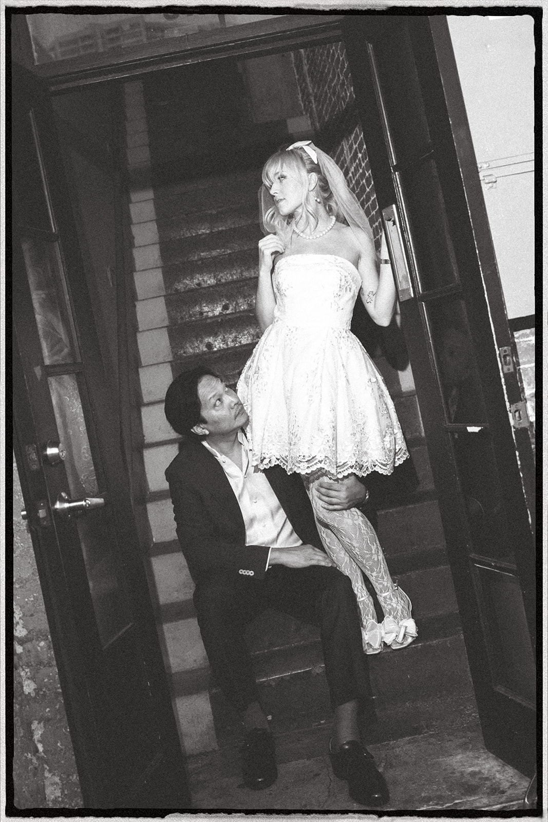 A playful moment between a bride and groom, with the bride in a short white dress, sitting on a staircase, and the groom looking up at her lovingly.