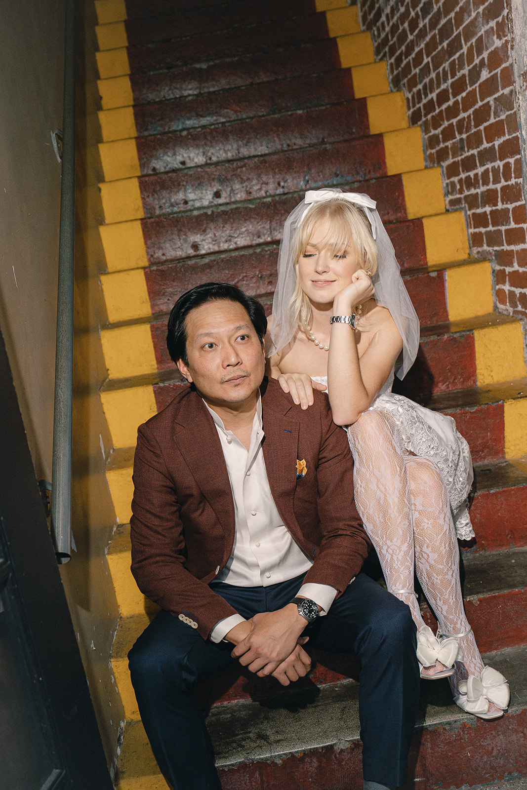 A joyful moment of a couple sitting on the stairs of an industrial building, with the woman in a wedding dress leaning on the man as they share a laugh, surrounded by a vibrant urban background.