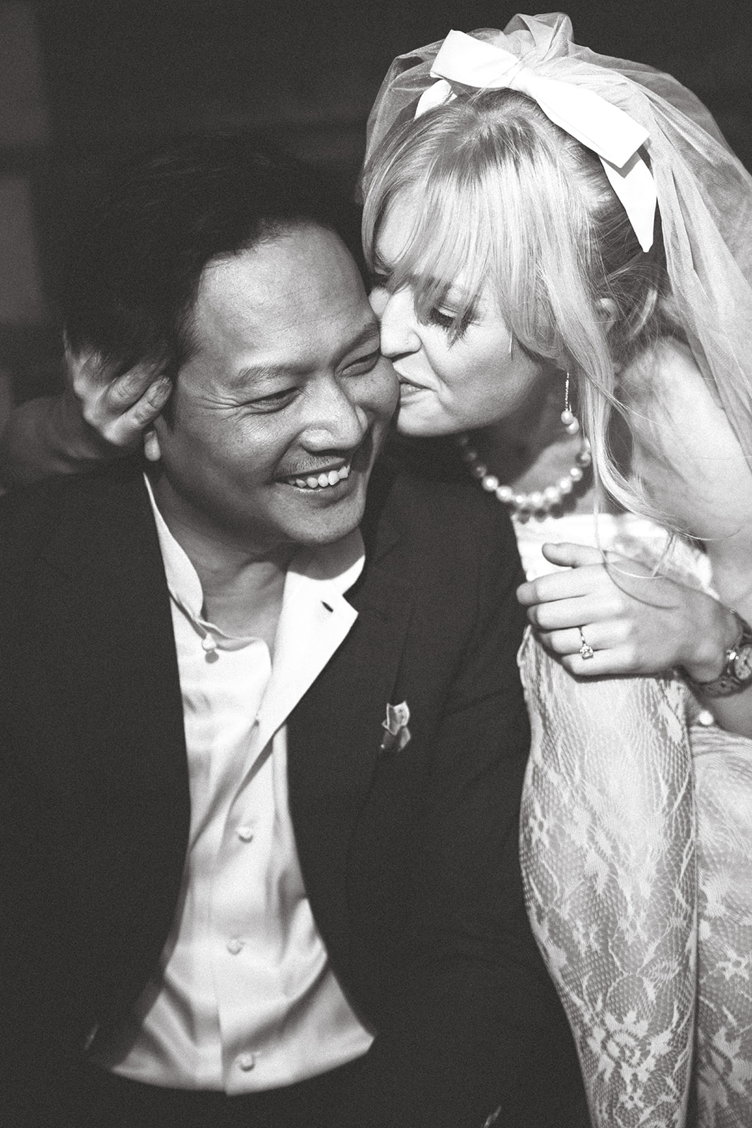 A candid black-and-white portrait of a couple during a photo session, with the bride playfully kissing her partner on the cheek while he laughs, capturing a joyful and intimate moment.