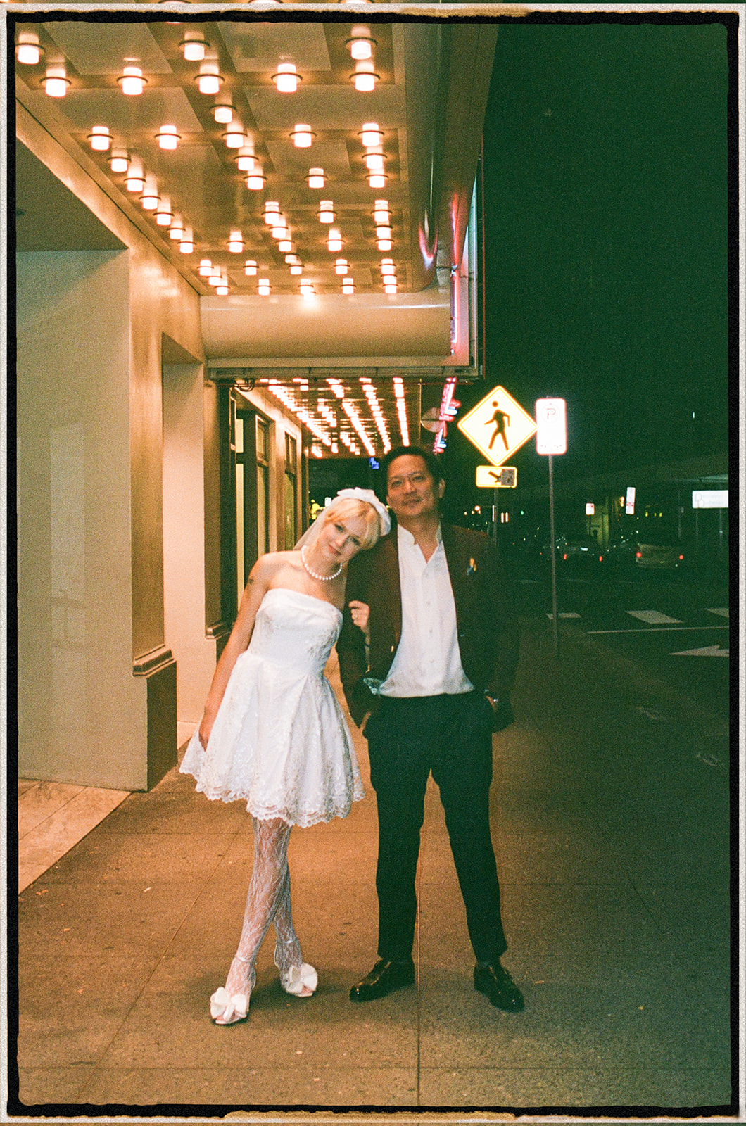 A couple standing together under a marquee sign, with the woman in a short white dress and lace stockings, and the man in a dark suit, with city lights glowing above them at night.