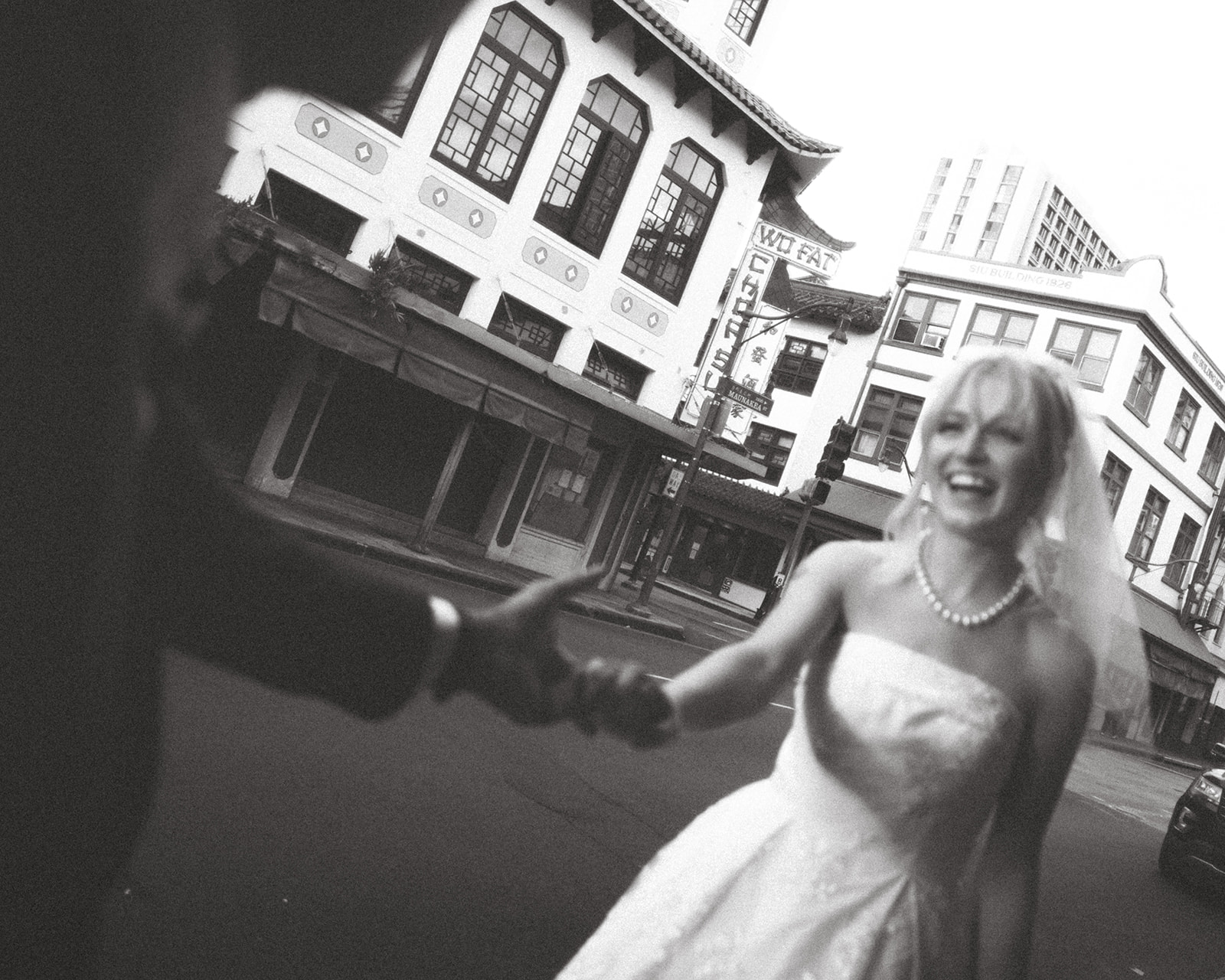  A joyful bride and groom hold hands and laugh in front of the Wo Fat sign in Chinatown, Honolulu, creating an energetic and carefree atmosphere.