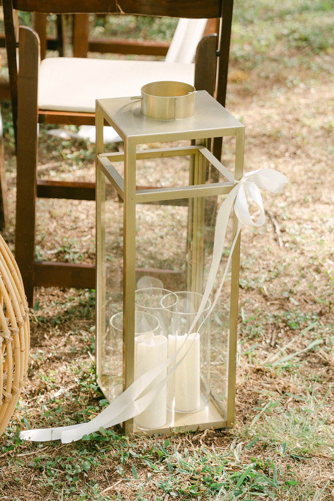 A gold-framed lantern with glass panels sits on the grass, adorned with a white ribbon tied in a delicate bow. Inside, a set of white pillar candles adds a soft, romantic touch to the outdoor wedding décor.