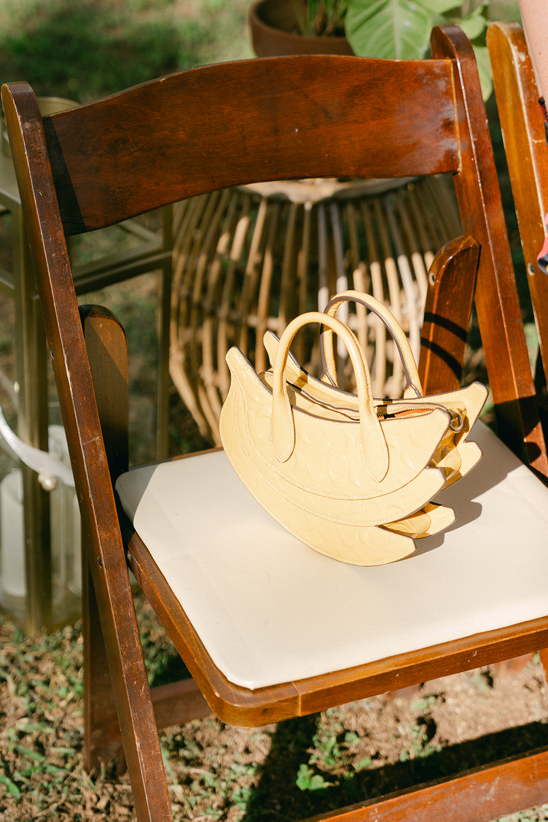 A stylish yellow handbag with a unique curved design rests on a wooden wedding chair. The warm tones and natural lighting add to the effortless, elegant aesthetic of the outdoor wedding setup.