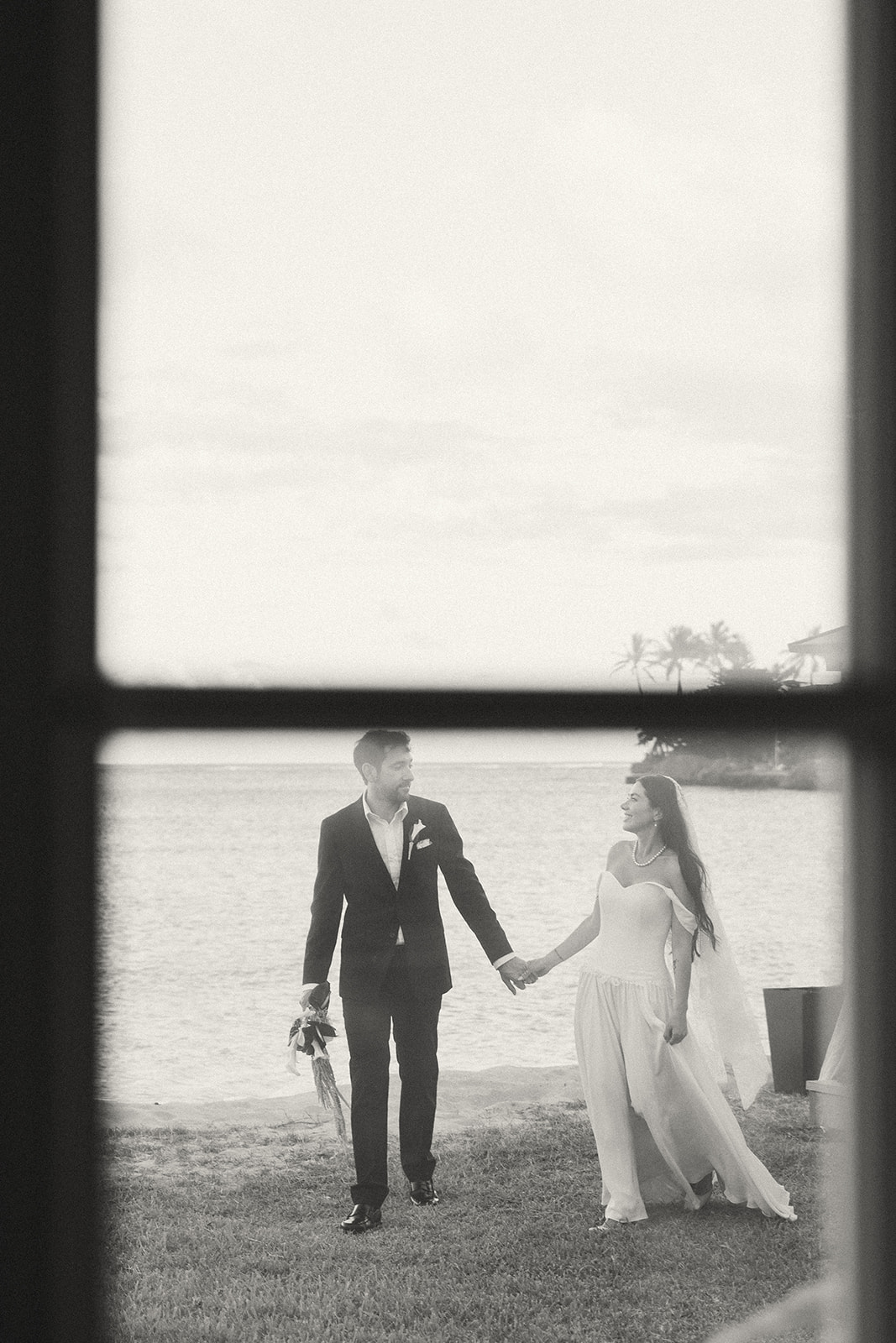 A couple strolling hand-in-hand by the ocean, captured through a window frame for a candid, intimate feel