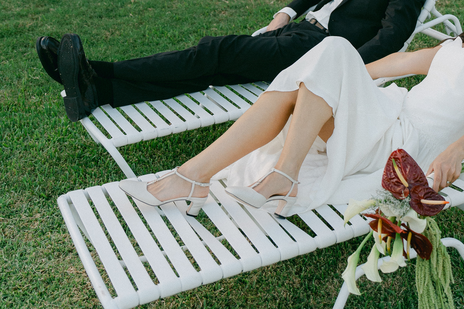A laid-back moment of a bride and groom lounging on the grass, her white heels and vibrant bouquet stealing the scene.