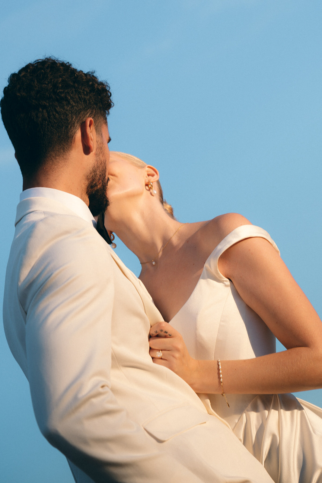 A bride in an elegant off-the-shoulder gown leans in to kiss her groom, who is dressed in a light suit, with the golden sunlight casting a dreamy glow against a clear blue sky.