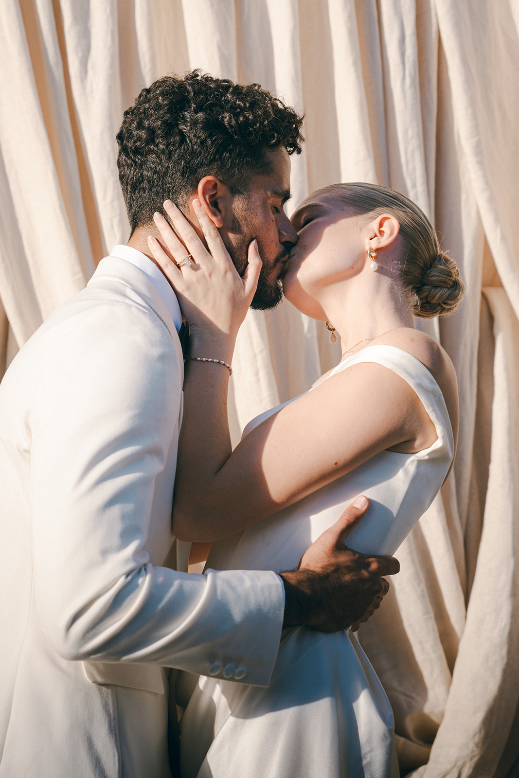 A newlywed couple sharing a passionate kiss in warm sunlight, both dressed in elegant white attire against a draped fabric backdrop.