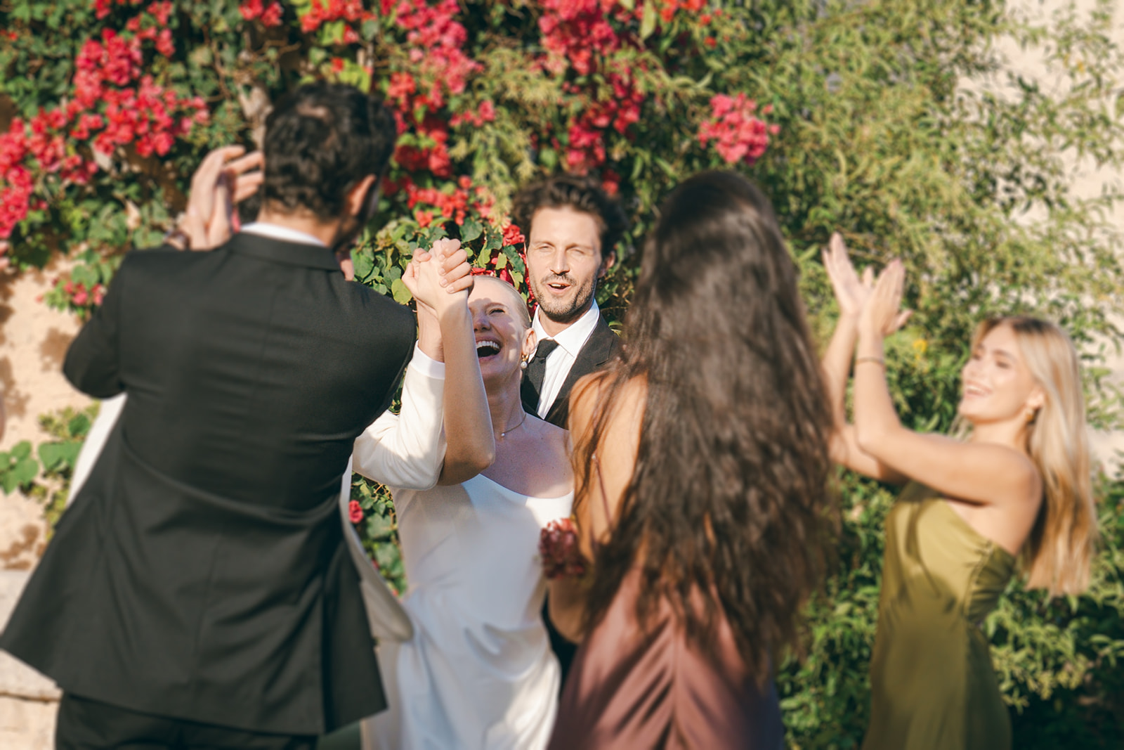 A joyful moment of a bride dancing with guests outdoors, surrounded by colorful flowers and greenery.
