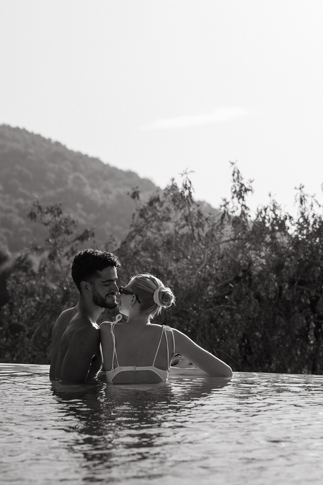 A couple enjoying a serene moment in an infinity pool, facing each other and smiling, with a beautiful mountainous backdrop.