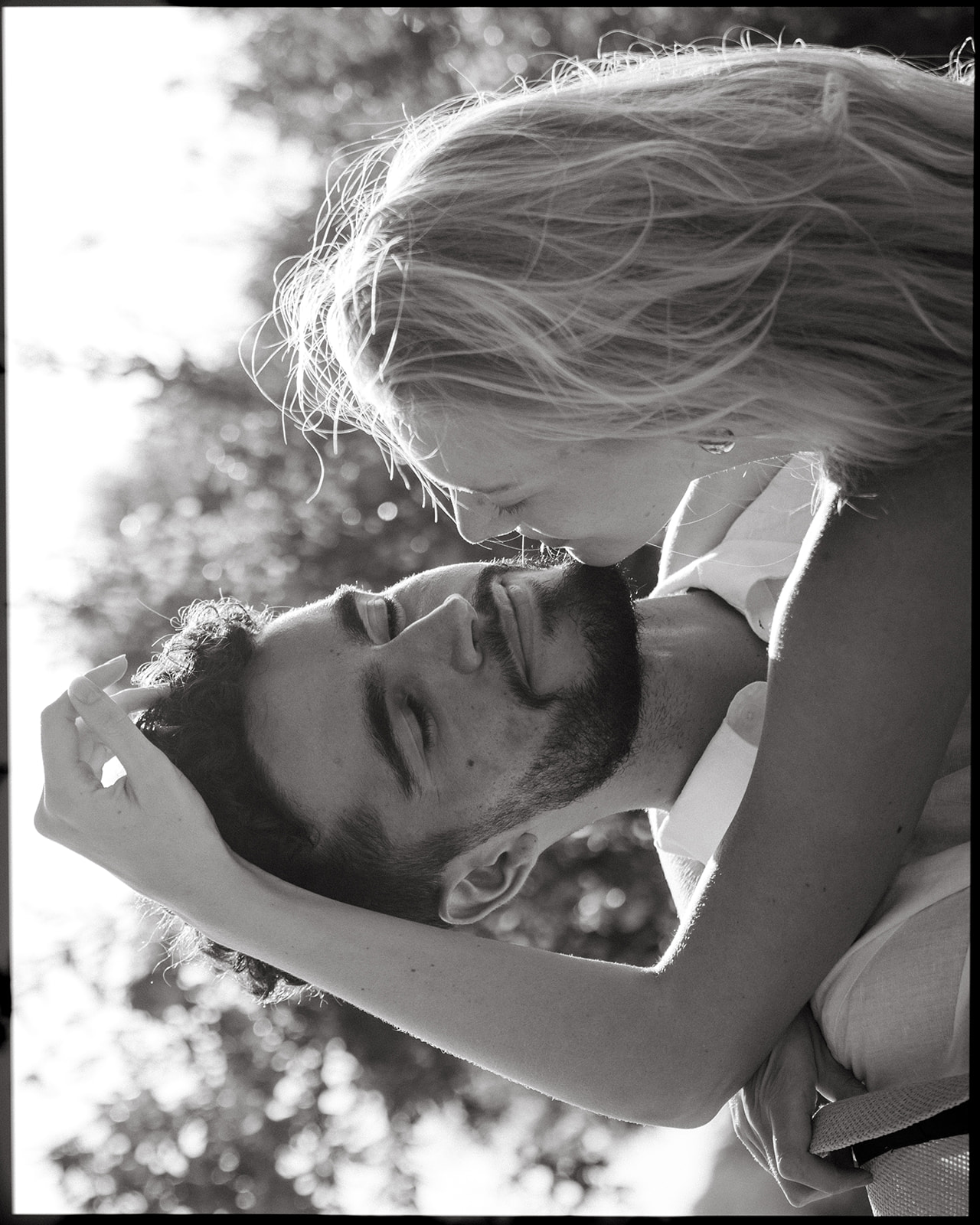 A black-and-white portrait of a couple sharing a tender moment, with their faces close together and the woman resting her hand gently on the man's head as they smile at each other in a sunlit outdoor setting.