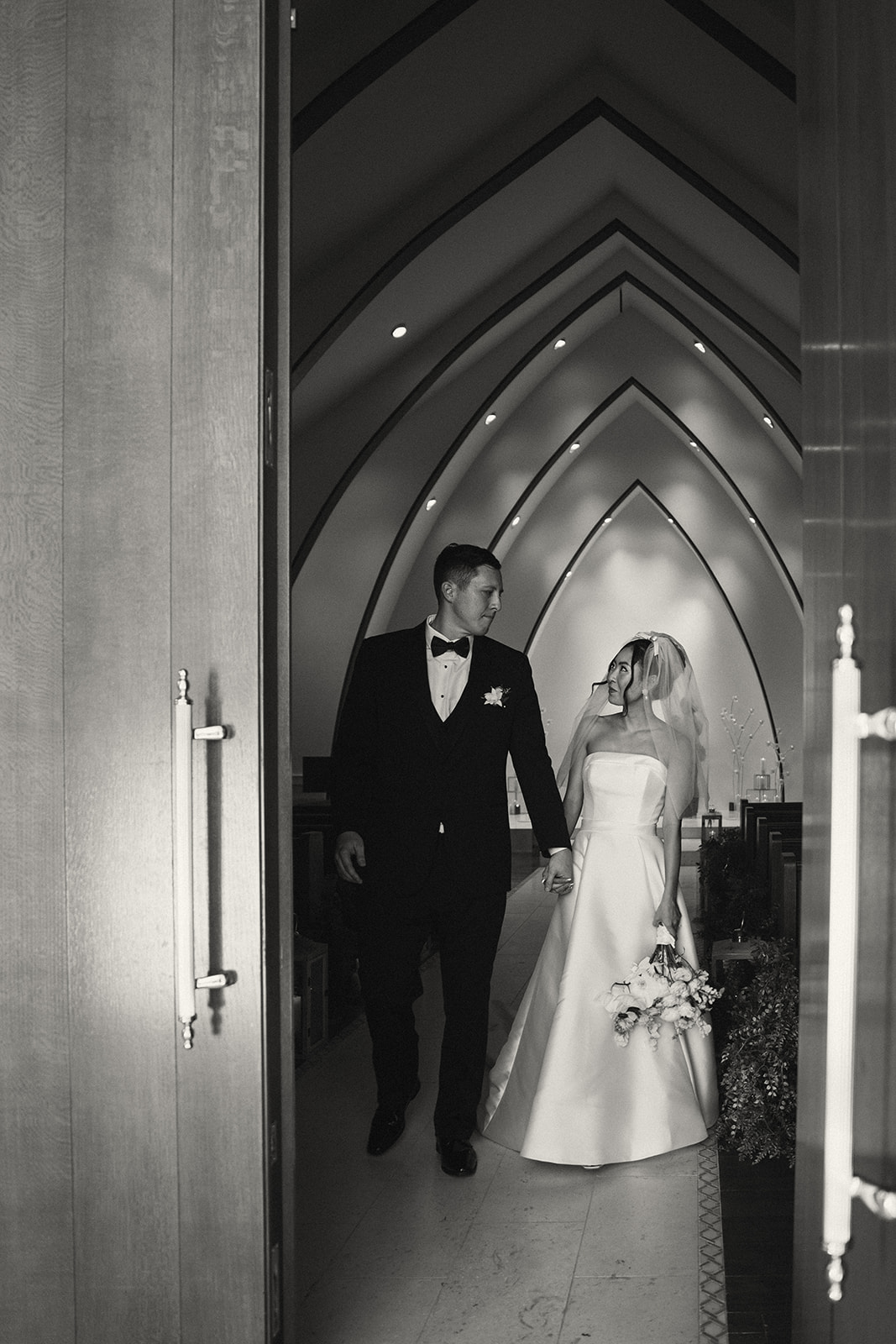 A black and white photo of a newlywed couple exiting the chapel, hand in hand, with a joyful expression as they walk through large wooden doors.