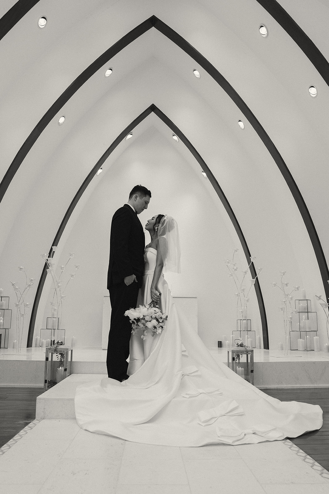 A black and white portrait of a bride and groom standing on the altar, gazing at each other lovingly, with the chapel's dramatic arched architecture framing them.