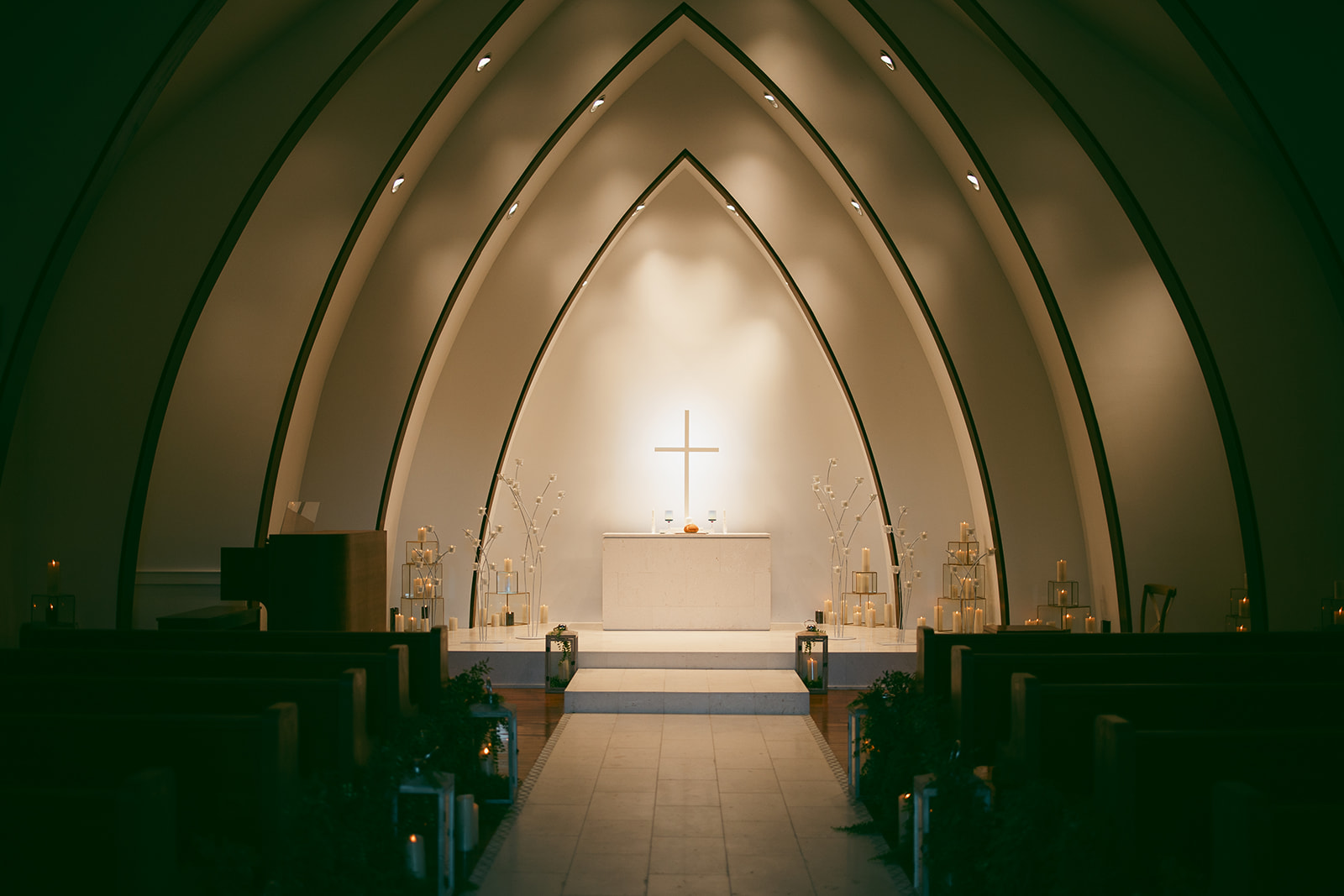 A modern chapel interior with arched ceilings and a softly illuminated altar featuring a cross, candles, and elegant floral arrangements.