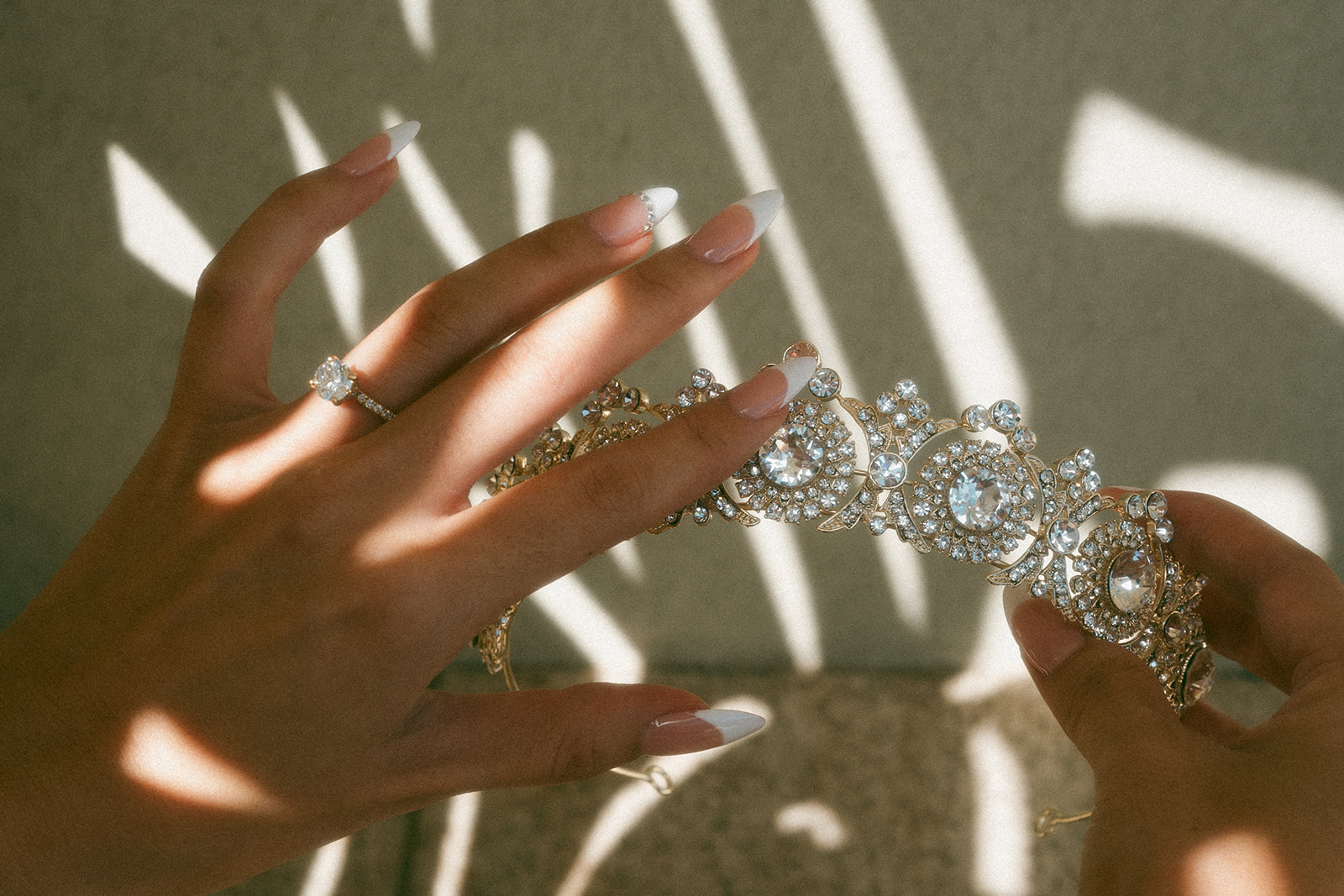 A close-up of a bride’s hands holding an ornate crystal tiara, with sunlight casting dramatic shadows through delicate lines. A sparkling engagement ring and perfectly manicured nails add to the elegant composition.
