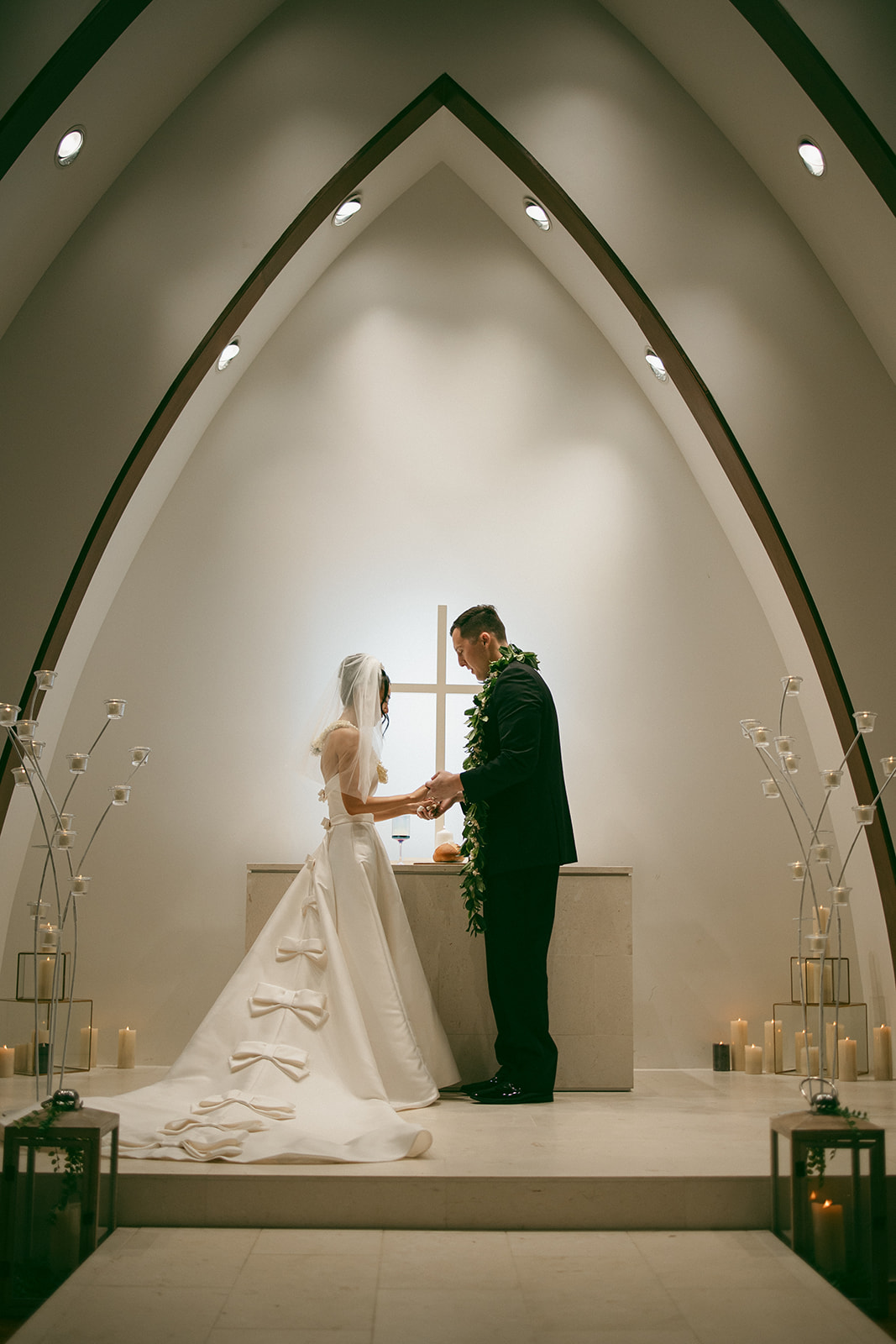 A bride and groom standing at the altar, exchanging vows in front of a softly glowing cross, with delicate candles and floral arrangements surrounding them.