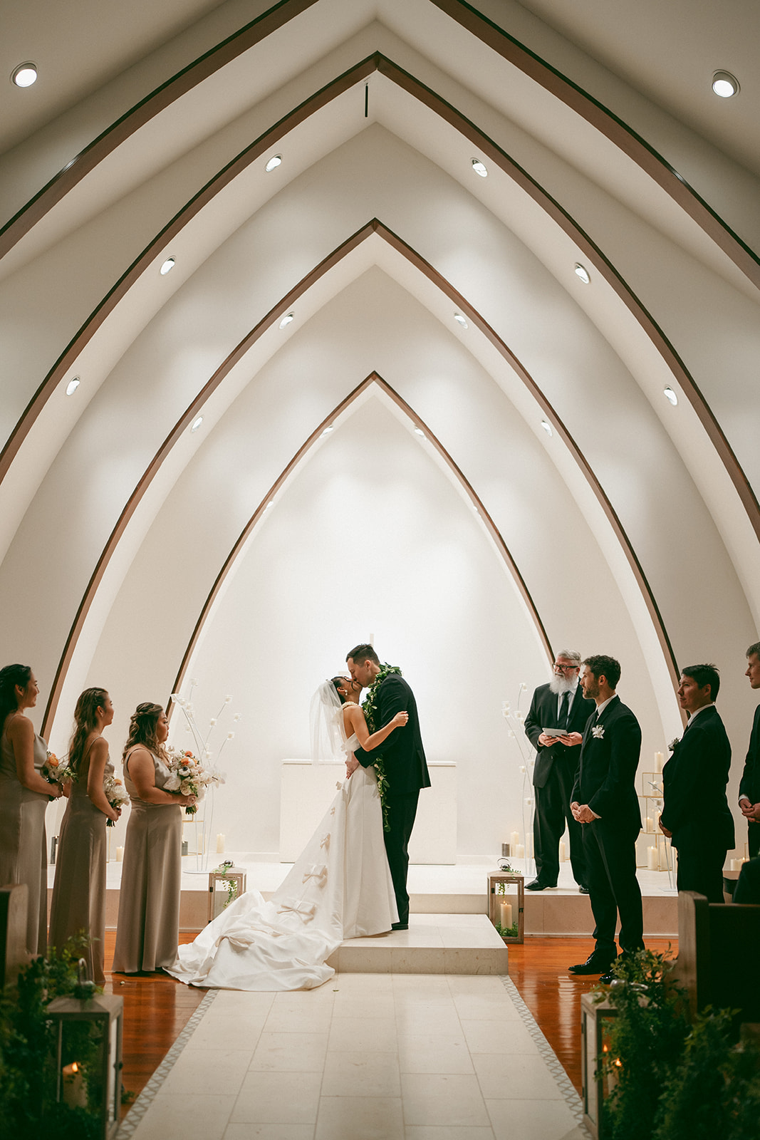 A bride and groom sharing their first kiss as newlyweds in a beautifully lit chapel, with their wedding party standing beside them.