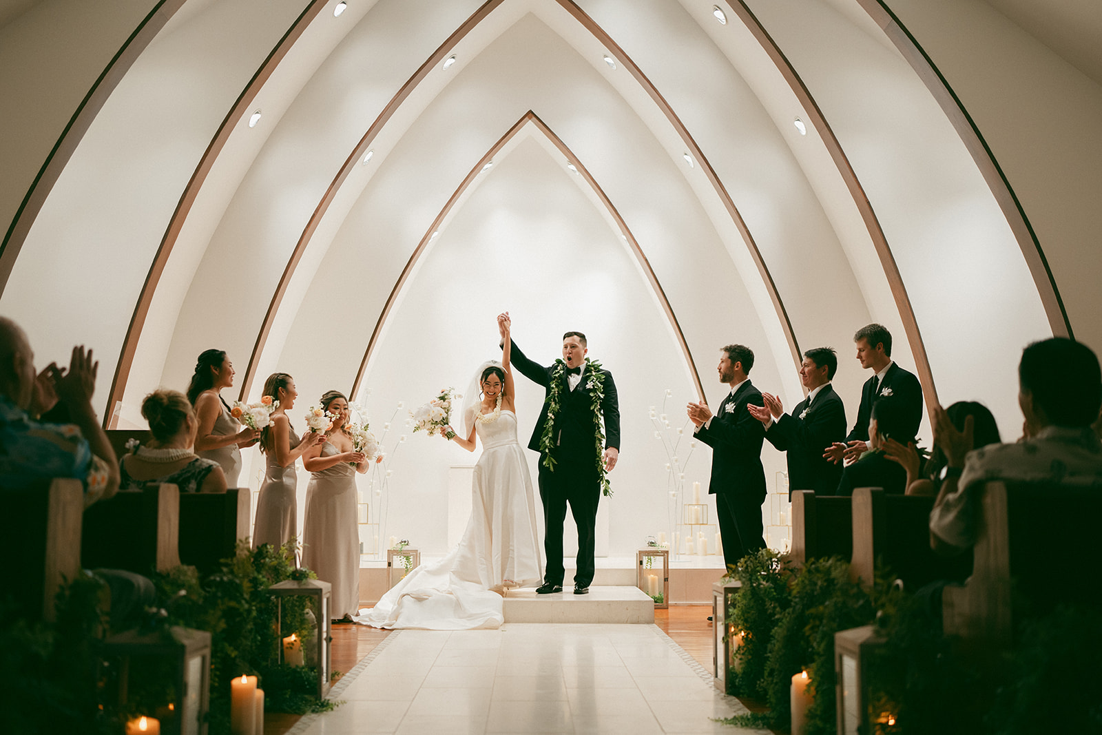 A couple stands at the altar, raising their hands in celebration after exchanging vows, surrounded by their wedding party in an elegant chapel.