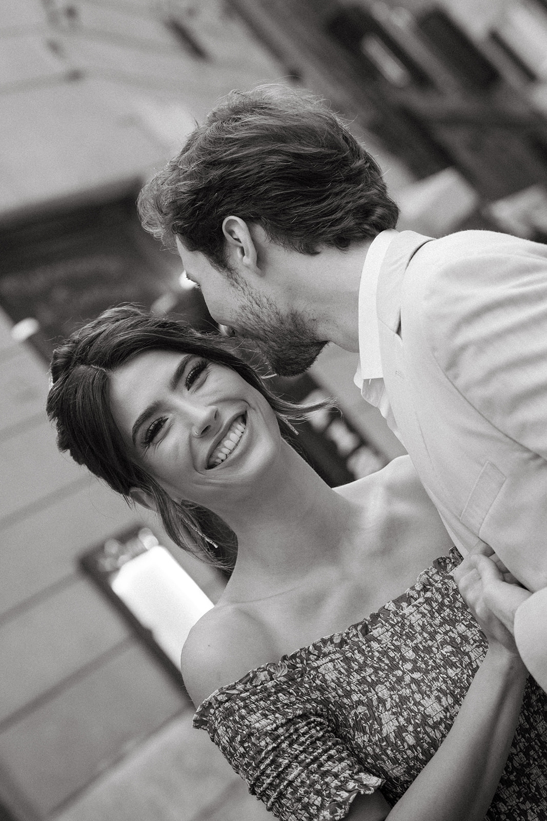 A black and white image of a smiling bride with tousled hair as her groom whispers something to her.