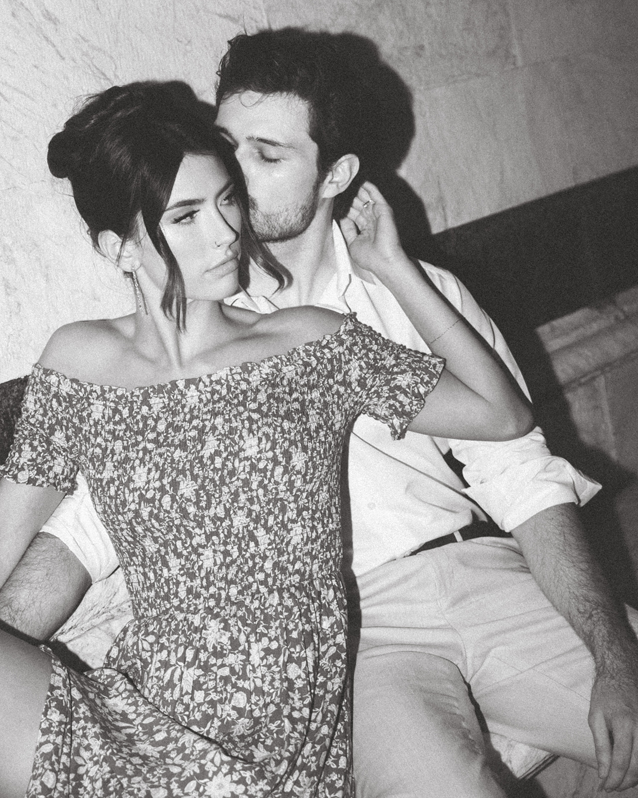  A black and white intimate portrait of a couple sitting closely against a textured wall. The woman, wearing an off-the-shoulder floral dress, gazes away while the man leans in, pressing a soft kiss to her temple. The image captures a quiet, romantic moment with a timeless feel.
