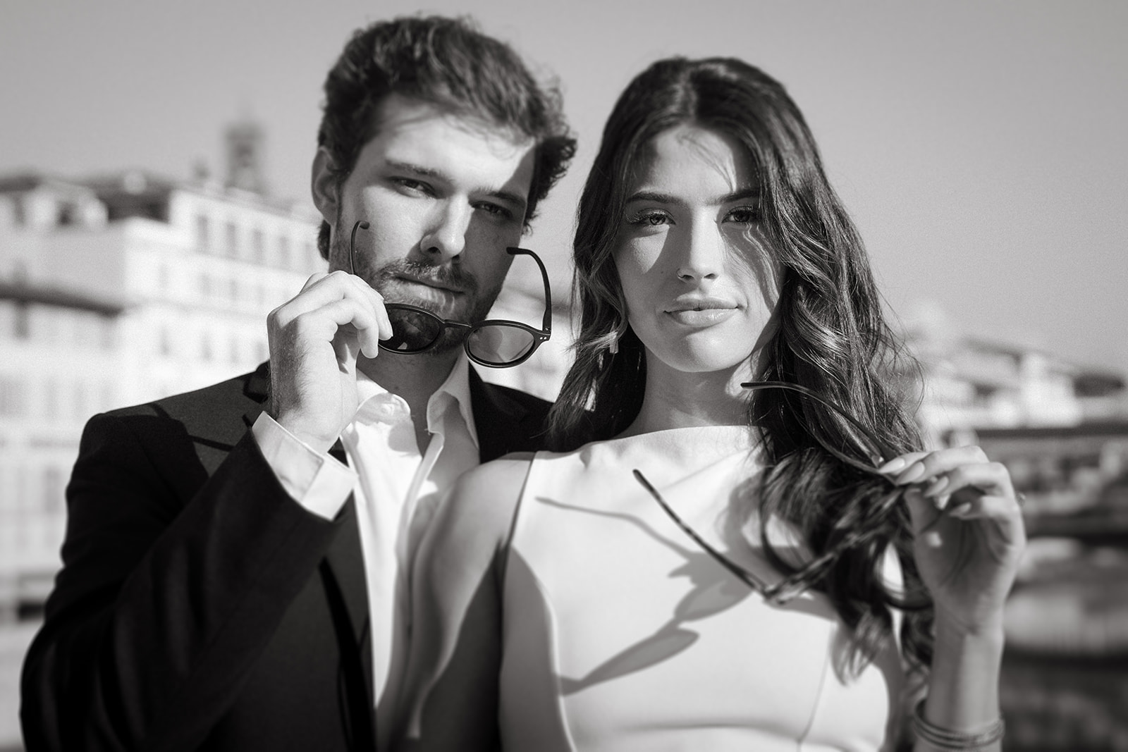 A black-and-white portrait of a man and a woman standing close together, both holding sunglasses. The man is looking at the camera, while the woman smiles softly, and they are dressed in formal attire against an urban backdrop.