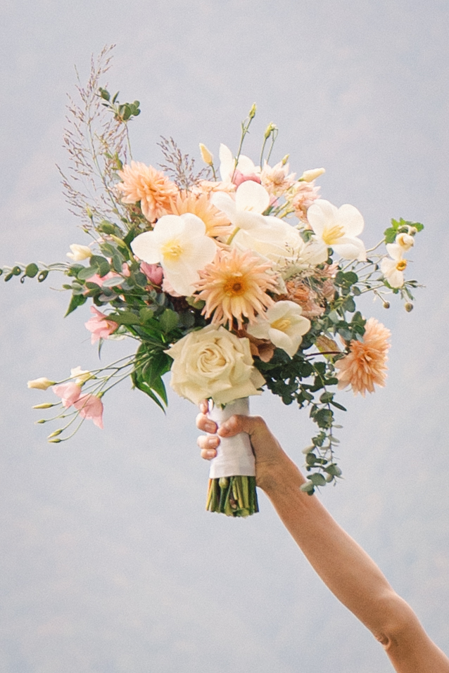 A beautifully arranged bridal bouquet featuring soft peach and white flowers, accented with lush greenery, held high against a blurred natural backdrop.
