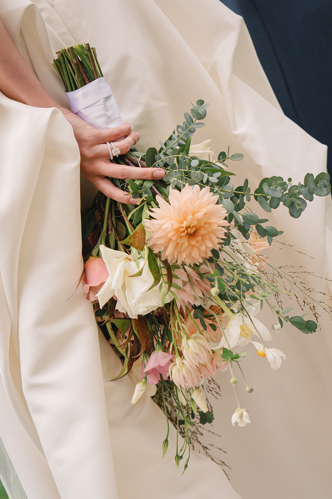 A close-up of the bride's hand holding a bouquet, showcasing her engagement ring as the flowers blend with her dress.