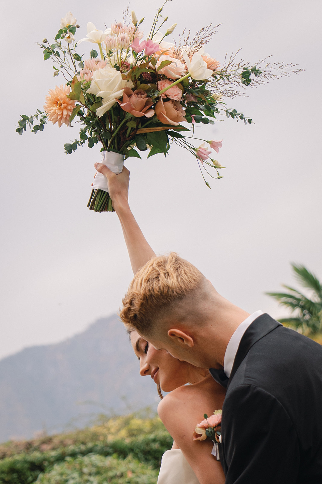 A couple celebrating with the bride holding her bouquet high in the air, while her partner embraces her from behind.