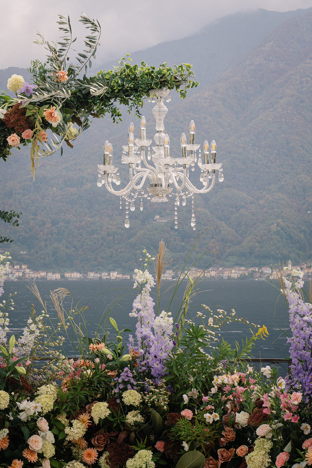 A wedding ceremony setup with a chandelier hanging amidst an array of pastel flowers, overlooking a scenic mountain backdrop.