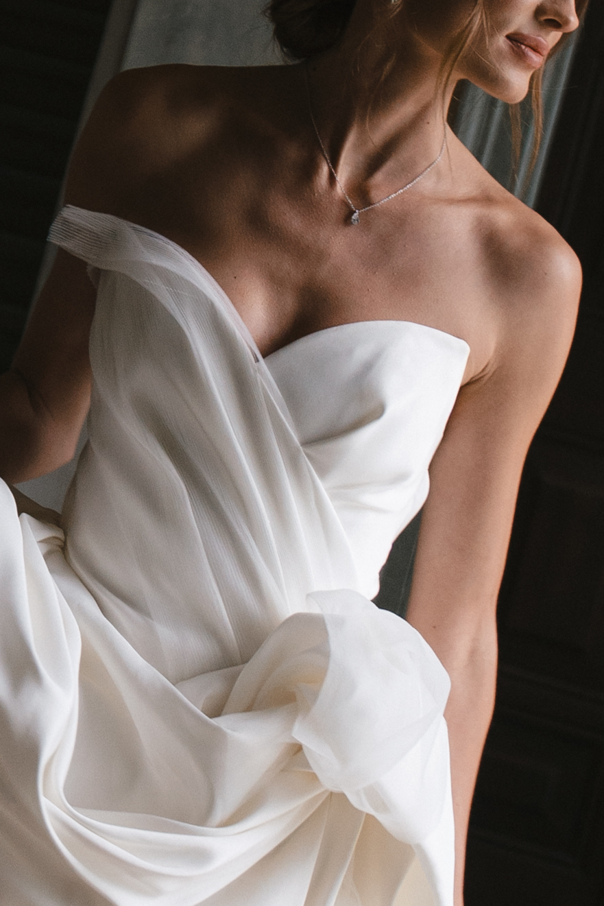 A close-up of a bride wearing a modern off-the-shoulder white wedding gown with delicate draping and a structured bodice.