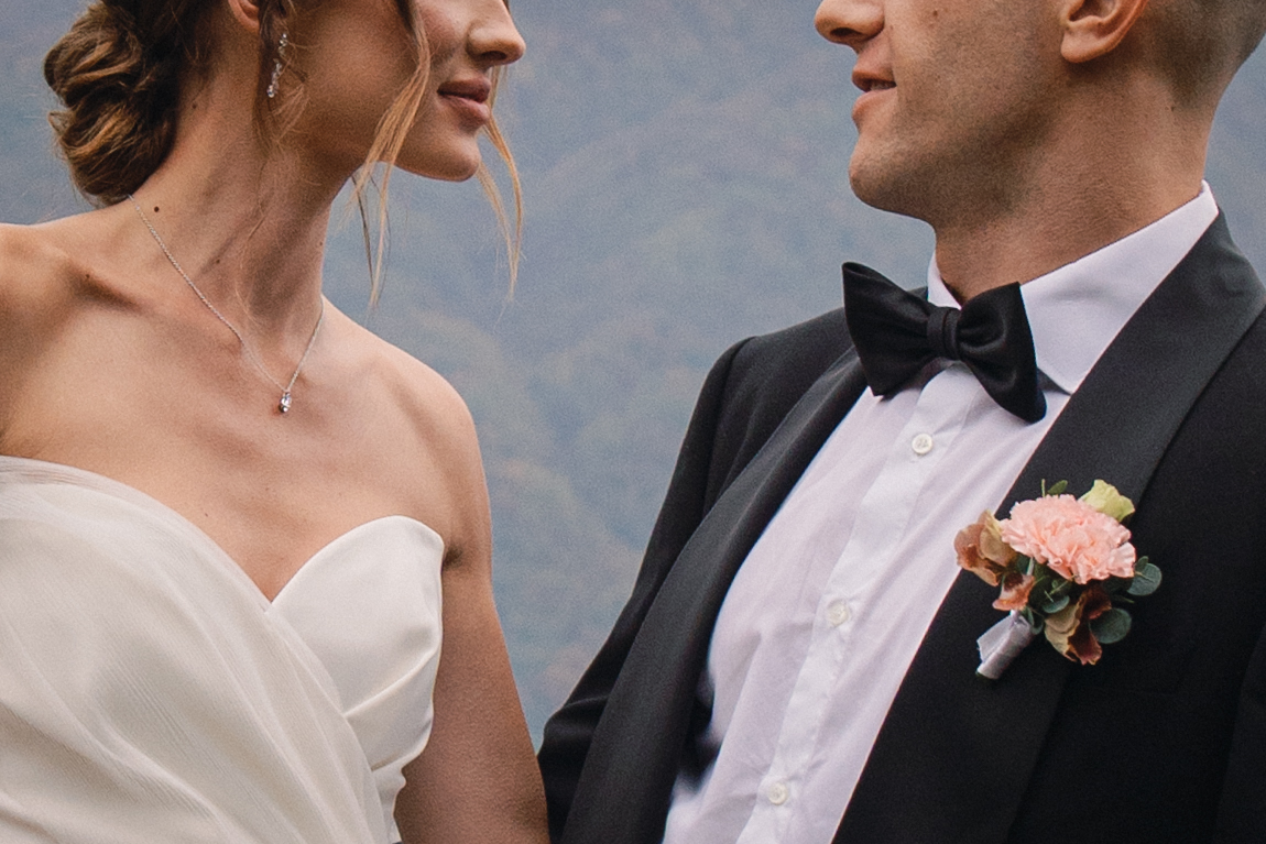 A close-up of a bride and groom facing each other, dressed in wedding attire, with soft mountain scenery in the background.