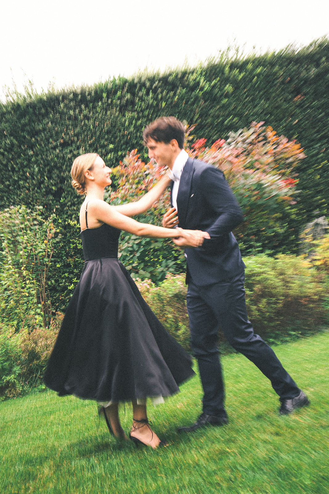 A stylishly dressed couple twirls on a lush green lawn, the woman’s black dress swaying with movement. Their playful expressions and joyful energy capture the romance of the moment.