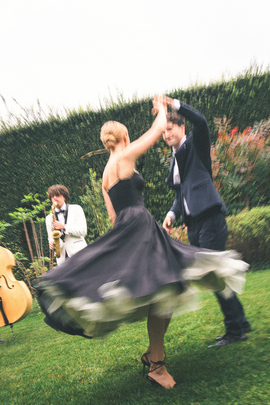 A woman in a vintage-inspired black dress spins gracefully as a man in a suit leads the dance. A saxophonist plays in the background, adding to the timeless elegance of the celebration.