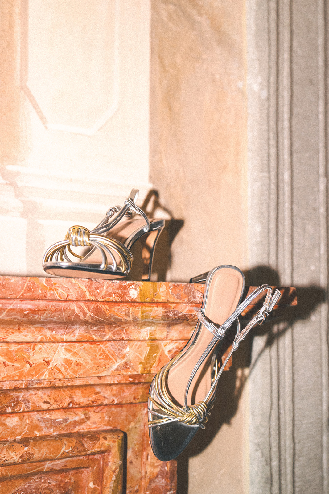 A stylish pair of metallic gold and silver strappy bridal heels placed on an ornate, warm-toned marble mantel.