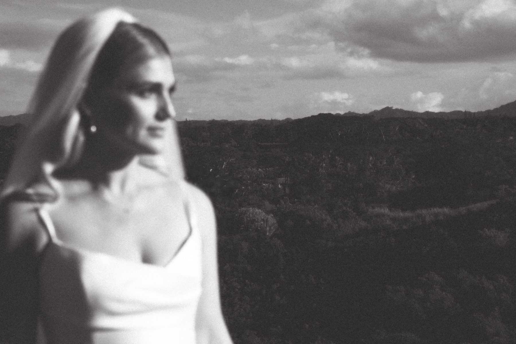A black-and-white photo of a bride in a simple white dress and veil, gazing off into the distance with a blurred mountainous landscape behind her, perfect for those seeking inspiration in a Photography Guide in Kapolei.