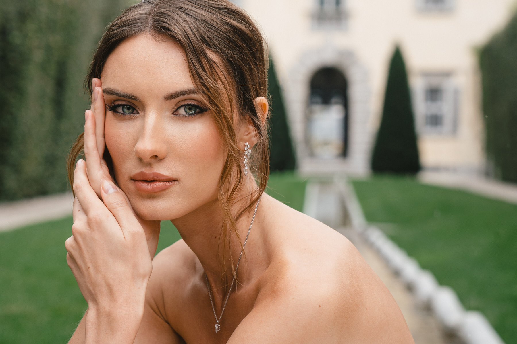 woman with brown hair in a strapless dress gently touching her face, standing in a garden with manicured hedges and a building in the background, showcasing a stunning example of Hawaii Bridal Photography.