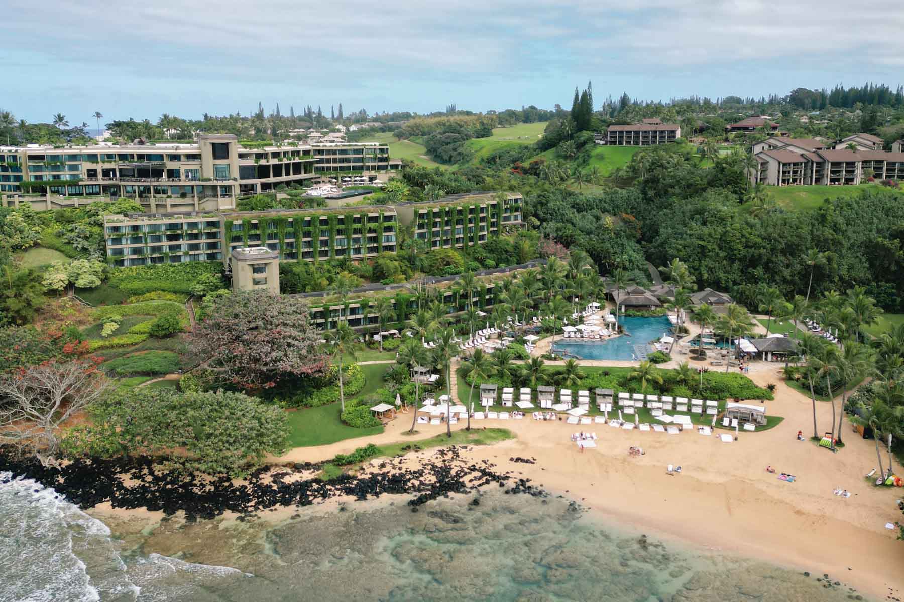 An aerial view of a luxury resort nestled on a cliffside overlooking the beach and ocean. The resort features modern architecture with sprawling green lawns and pools, perfect for relaxation and enjoyment.