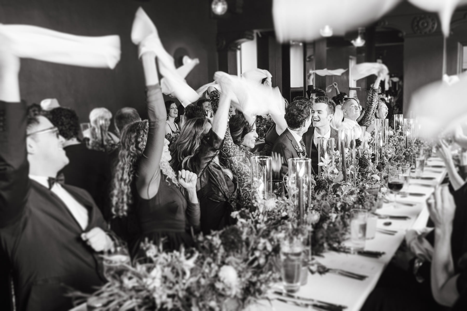 A joyful black-and-white image showing wedding guests waving cloth napkins in celebration at a reception table. People are dressed in formal attire, smiling, with floral arrangements on the table.
