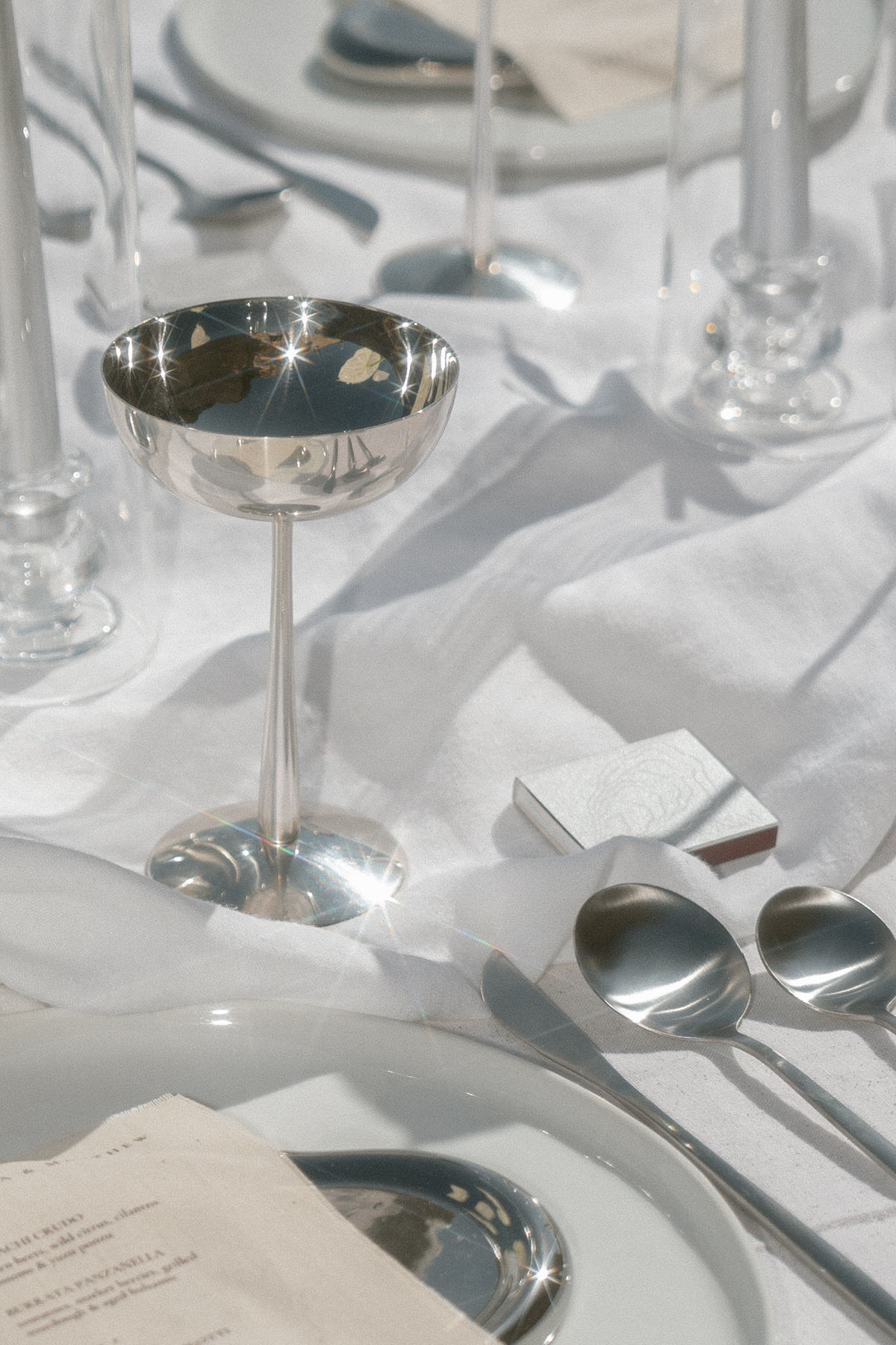 A table set for a wedding reception with sparkling silverware, a shimmering silver champagne glass, and decorative details like a folded napkin, resting on a pristine white cloth. The sunlight highlights the elegant and refined setting.