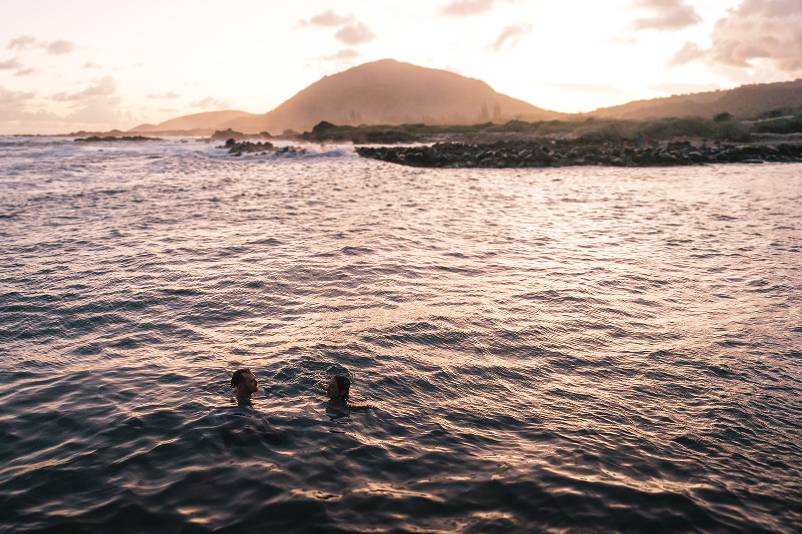 Couple swimming together in the ocean with the island's silhouette in the background, creating a serene and romantic scene in Marriage Proposal Photo Session in Oahu.