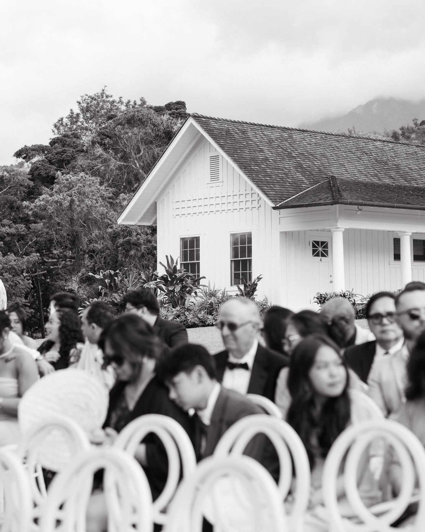 Black-and-white image of elegantly dressed guests seated outdoors by a charming white house, reflecting candid moments from a Hawaii ceremony in a Destination Wedding Photography Guide for Couples Planning a Hawaii Wedding.