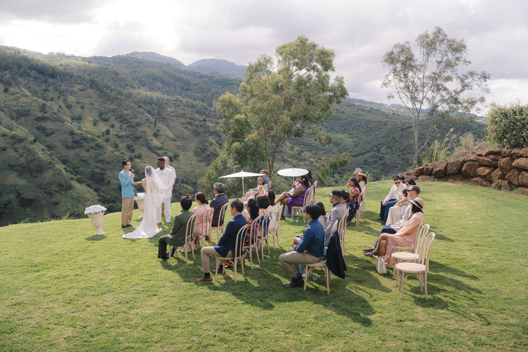 An intimate ceremony set on a grassy mountaintop in Hawaii, with guests seated and a couple exchanging vows beneath a cloudy sky—an unforgettable scene for a Destination Wedding Photography Guide for Couples Planning a Hawaii Wedding.